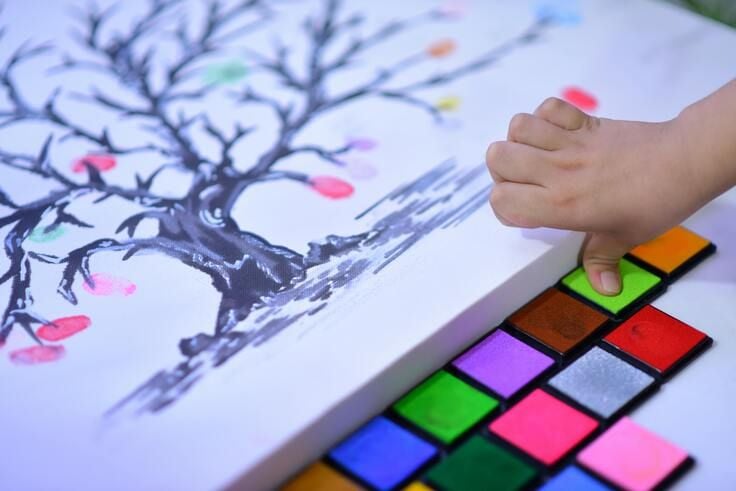 un tableau représentant un arbre, une palette de couleur et le doigts d'un enfant qui se frotte sur la couleur verte