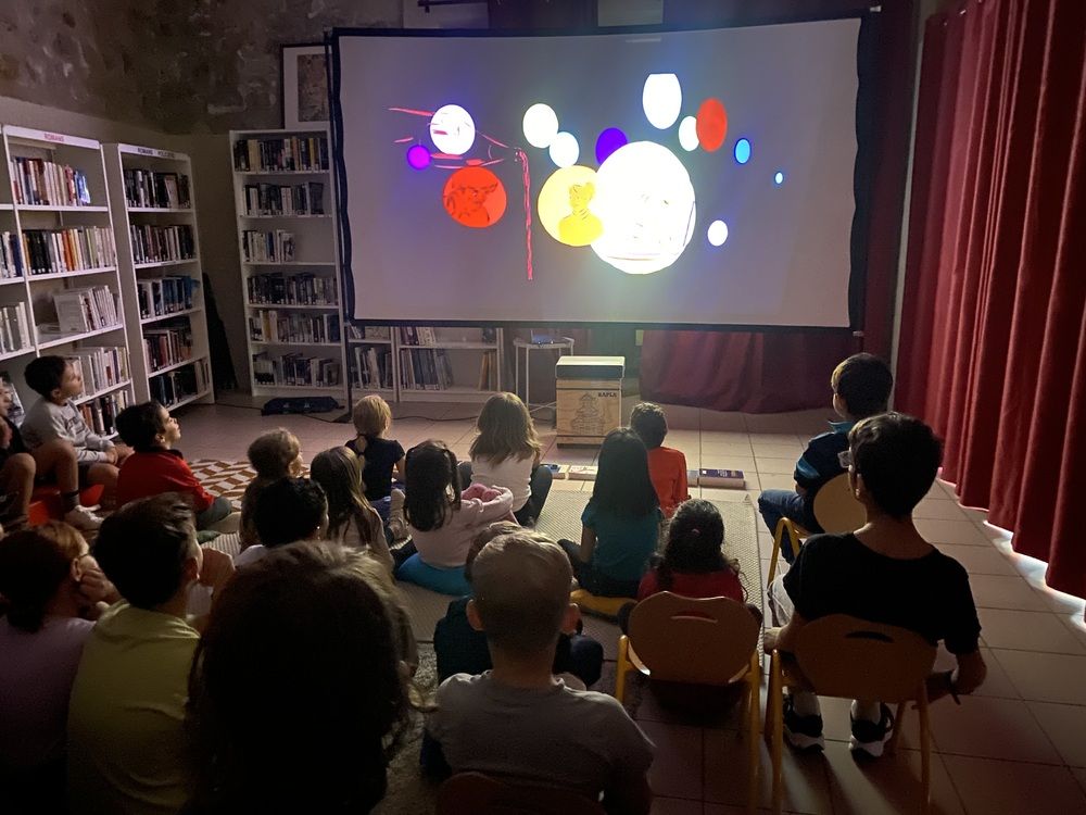 L'image représente des enfants dans une salle devant un grand écran où est projeté un dessin animé