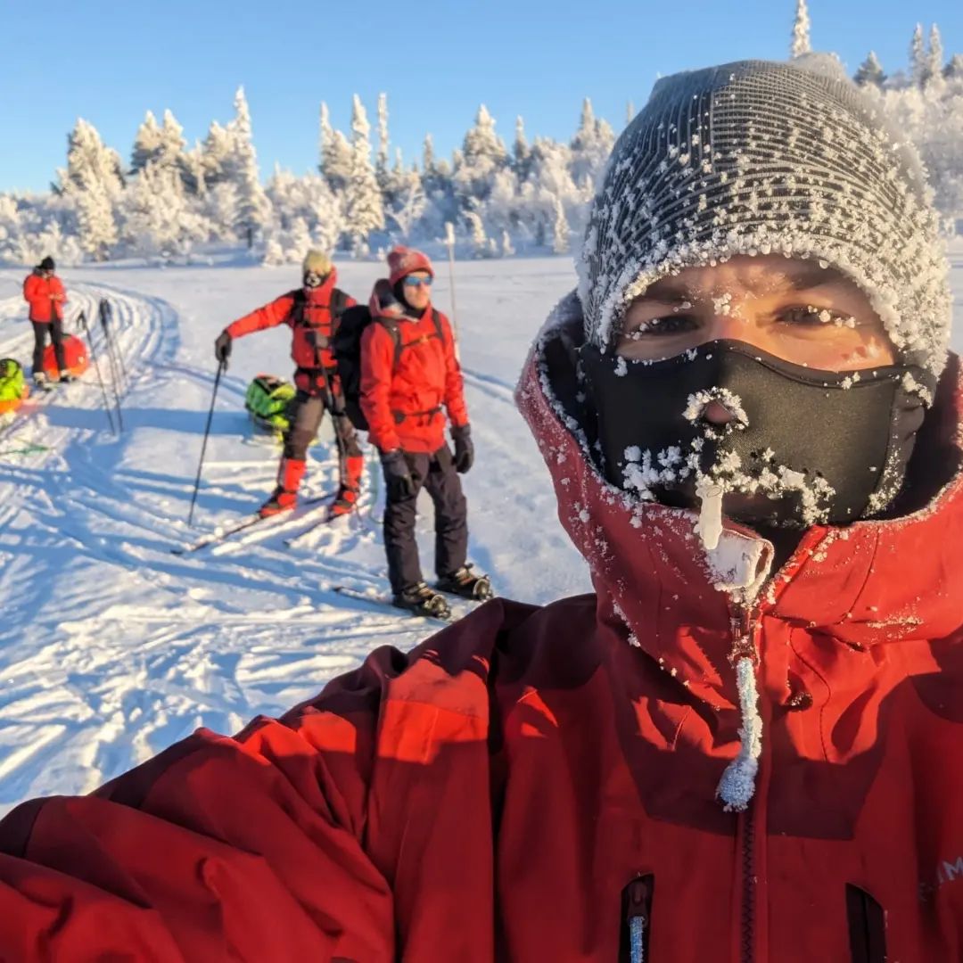 La photo représente un homme avec un bonnet, et des cristaux de glaces et de la neige dessus, on voit une piste de ski et des sapins enneigés